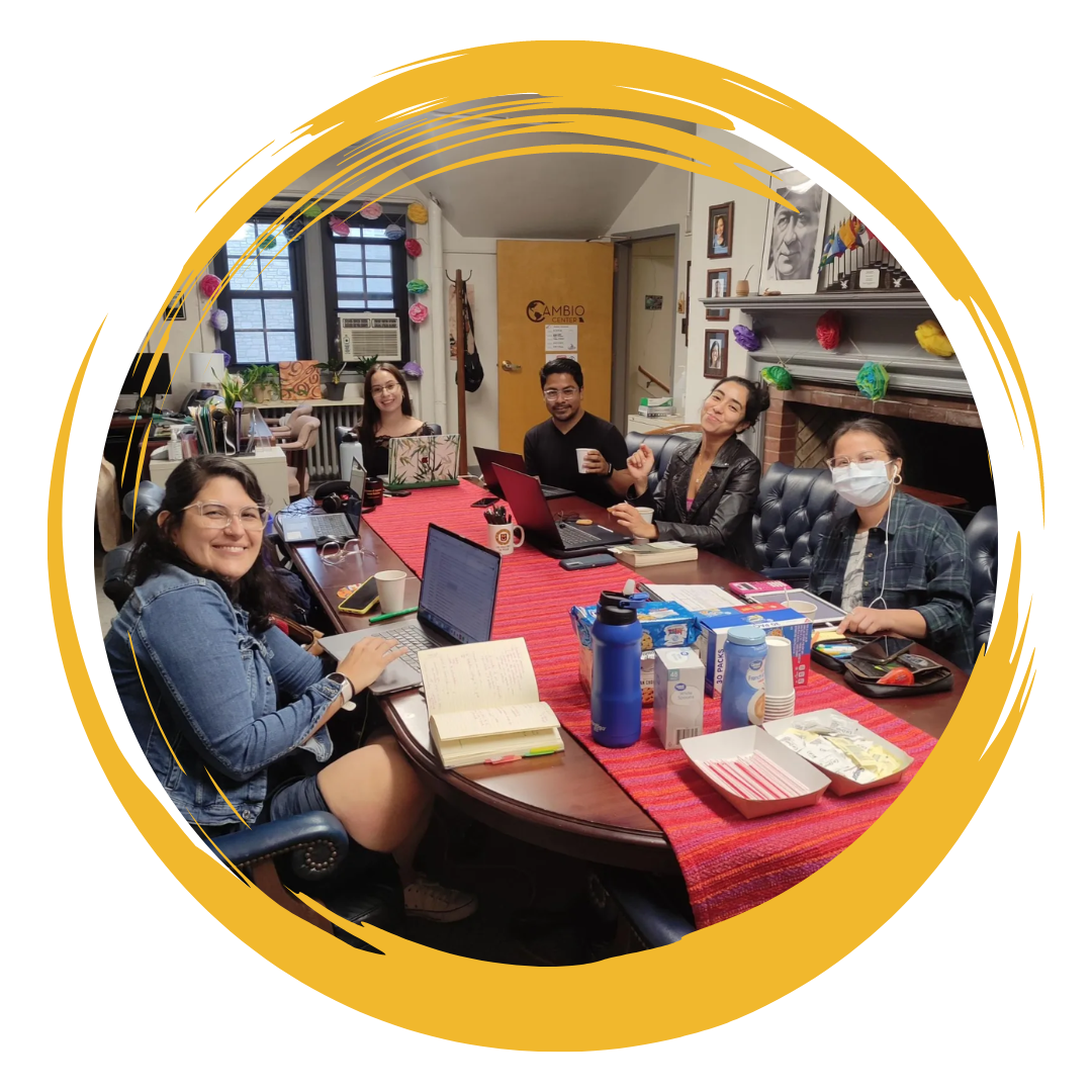 6 graduate students taking a break from studying to pose for a picture. There are 4 women smiling and one man while sitting in front of their laptops and books that are placed over a colorful fabric table runner. In the background, an office with a large picture of Cesar Chavez and beautiful large flowers made of paper. Image is framed by a gold circular accent.