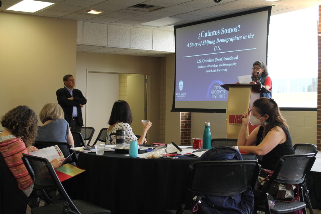 Image of Debora Bolton, presenting keynote speaker, Onésimo (Ness) Sandoval at the 20th Cambio de Colores Conference. In background, a screen as scene with powerpoint presentation that reads "¿Cuántos Somos? A Story of Shifting Demographics in the United States". People are sitting around tables listening and taking notes. 