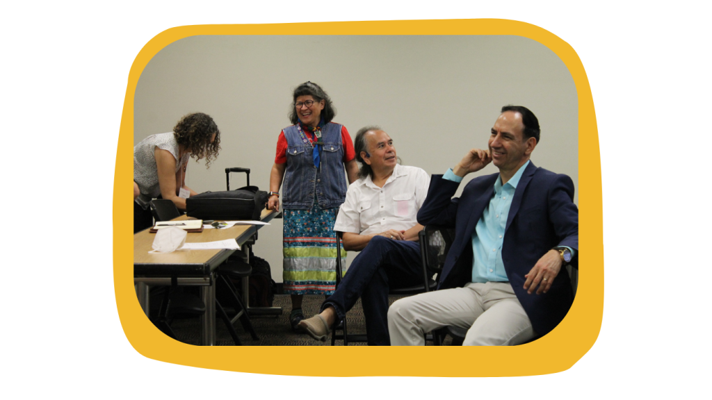 Image of 4 people laughing while discussing their research and projects at the 20th Cambio de Colores Conference. A woman stands at the center wearing an indigenous Ribbon skirt with hues of blue, green, and orange. Image is placed over a yellow uneven accent framing it.