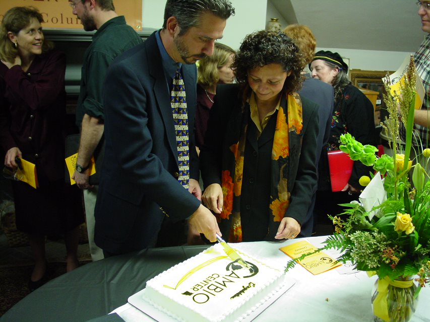 Image of Corinne Valdivia, Co-Founder of Cambio Center, cutting a cake for the Cambio Center opening. People are around them talking and flowers are in front of them. The cake they are cutting has white frosting with a logo image in black that reads "Cambio Center"